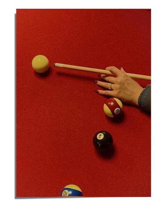 A red pool table with scattered billiard balls and a player's hand carefully positioning a shot with a cue stick. The lighting creates a warm and moody atmosphere, highlighting the texture of the felt and the contrast of the billiard balls.