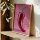 Framed artwork of a red chili pepper on a pink background, leaning against a wooden shelf.
