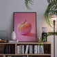 Framed artwork of a pink fruit on a shelf with books and decor items.