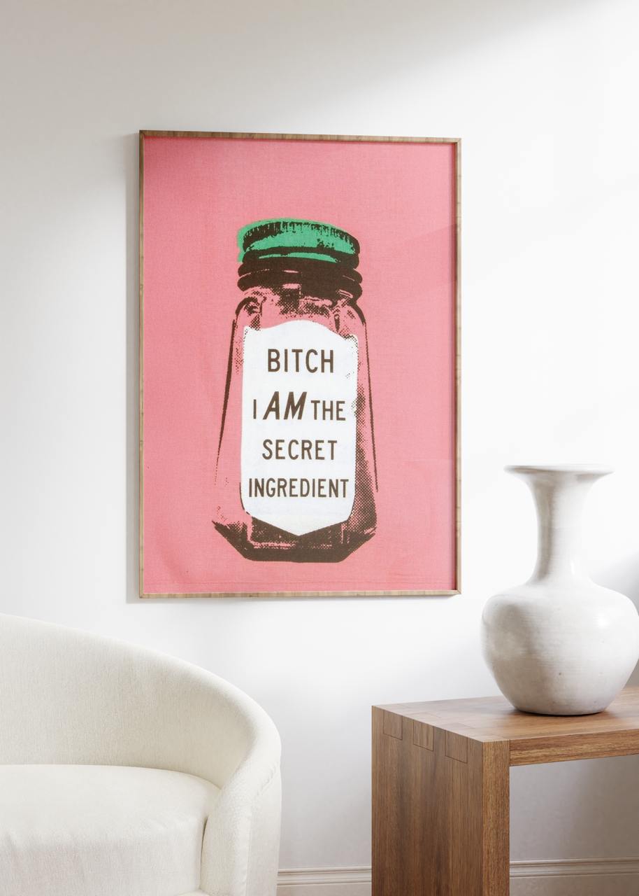 Framed artwork with a pink background and text on a wall above a white chair and wooden side table.
