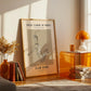 Framed artwork on a wall with a modern interior setting featuring a lamp and books.
