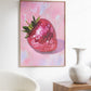 Framed artwork of a disco strawberry on a pink abstract background in a room with a chair and table.